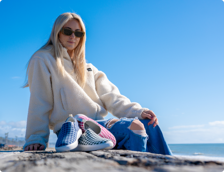 Woman in a cream fleece jacket and ripped jeans sitting on the beach, arranging various colorful Woolloomooloo sneakers on the sand