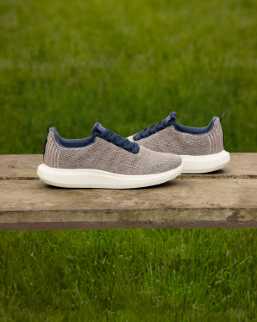 Pair of gray Woolloomooloo lace-up sneakers with white soles and navy blue accents, placed on a wooden bench with green grass in the background.