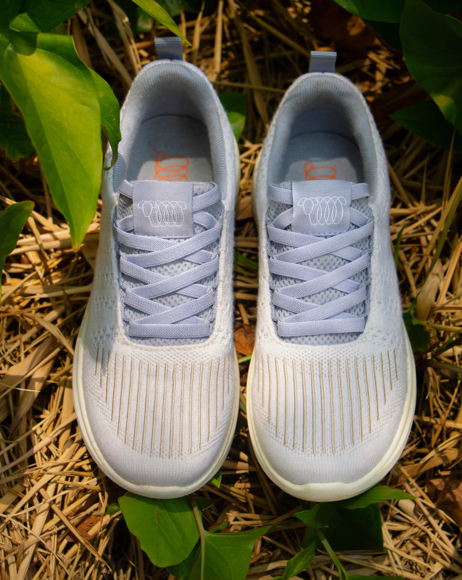 Pair of light gray Woolloomooloo lace-up sneakers with white soles, displayed outdoors on straw surrounded by green leaves