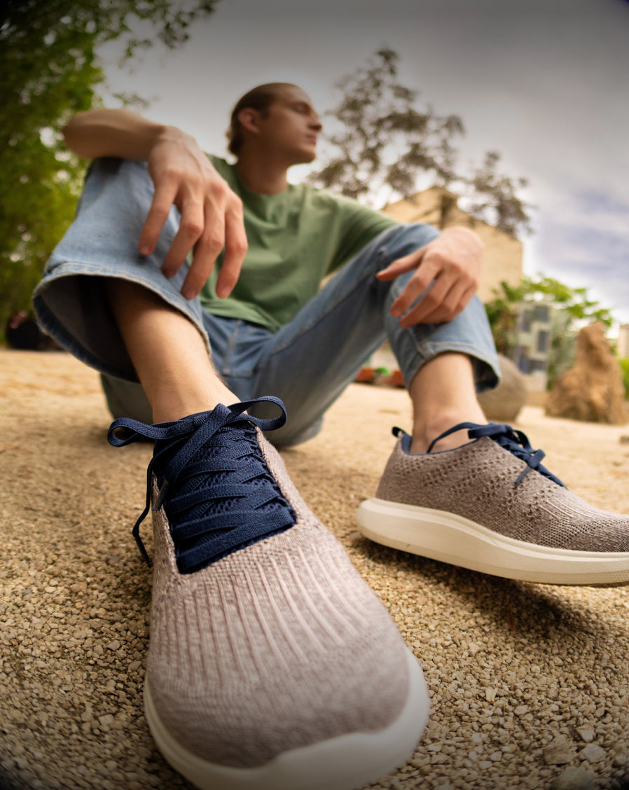 Man sitting on the ground outdoors wearing light brown Woolloomooloo sneakers with white soles and navy blue laces, dressed in a green shirt and light blue jeans